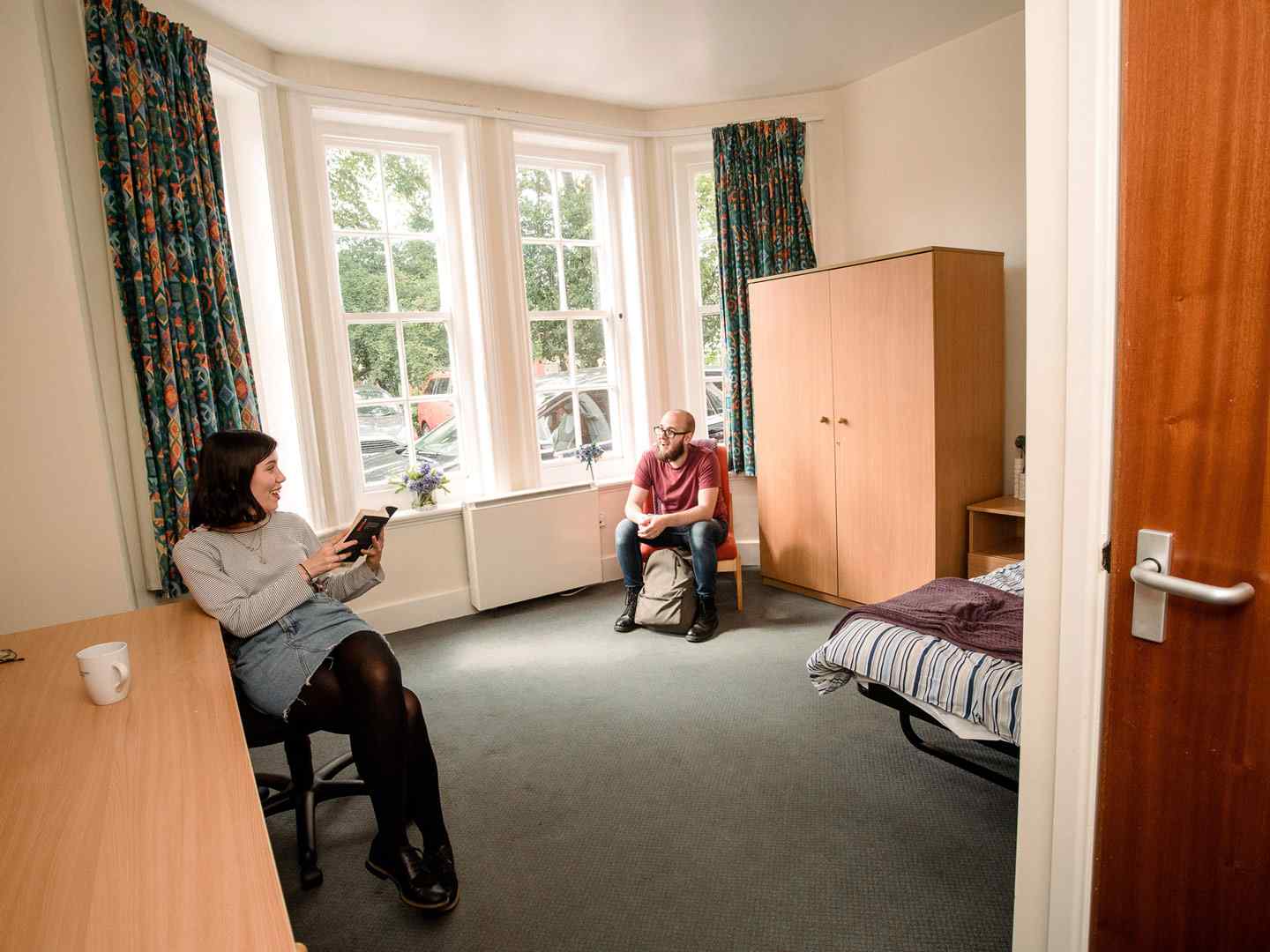 Two students sitting and chatting in a large ensuite room at City Residence. A large bay window is on the far wall, with a desk and wardrobe visible on either side. 