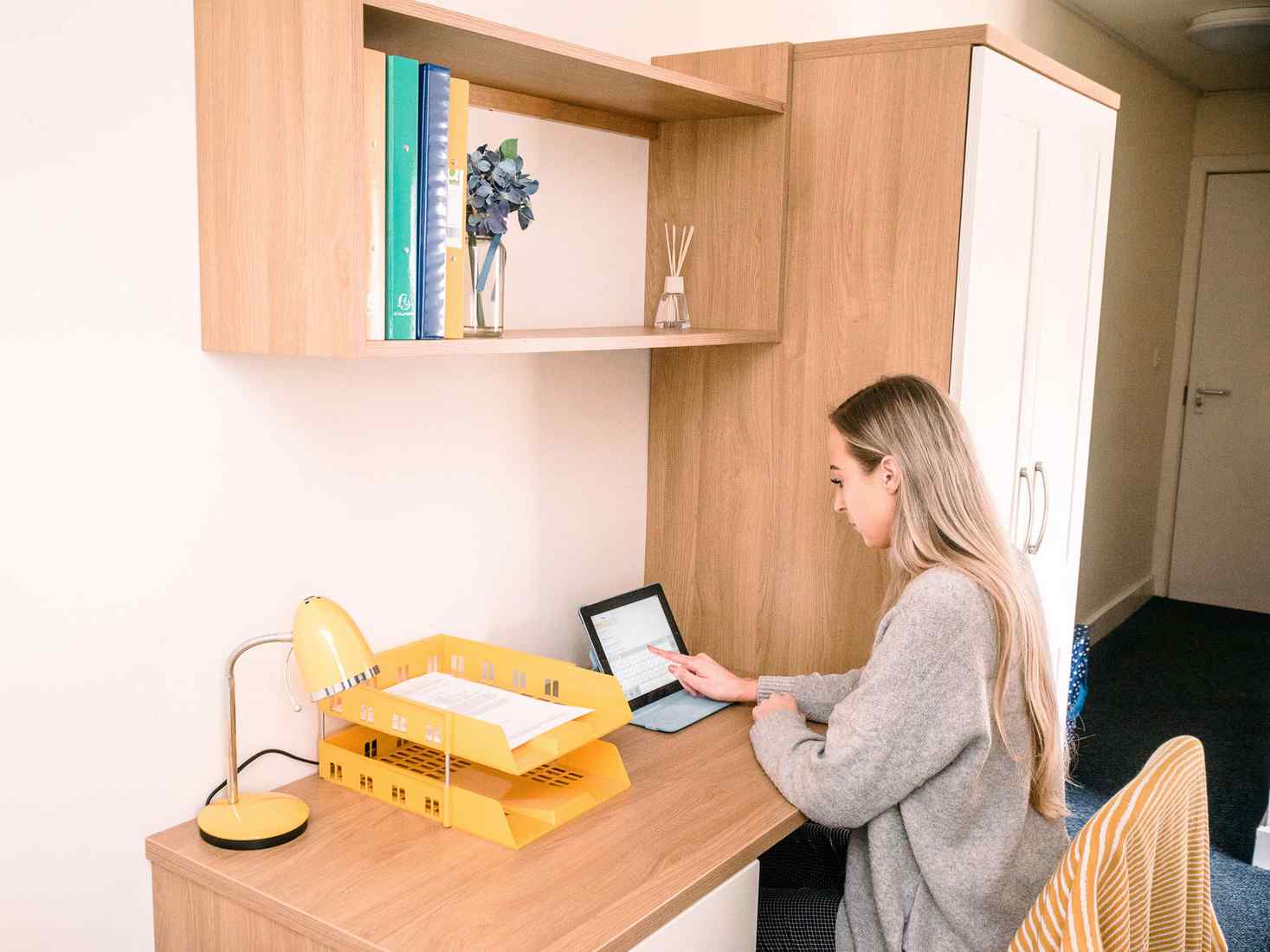 A student sits at a desk in a Garden Street standard ensuite room. There is a wardrobe next to the desk and shelving above. 