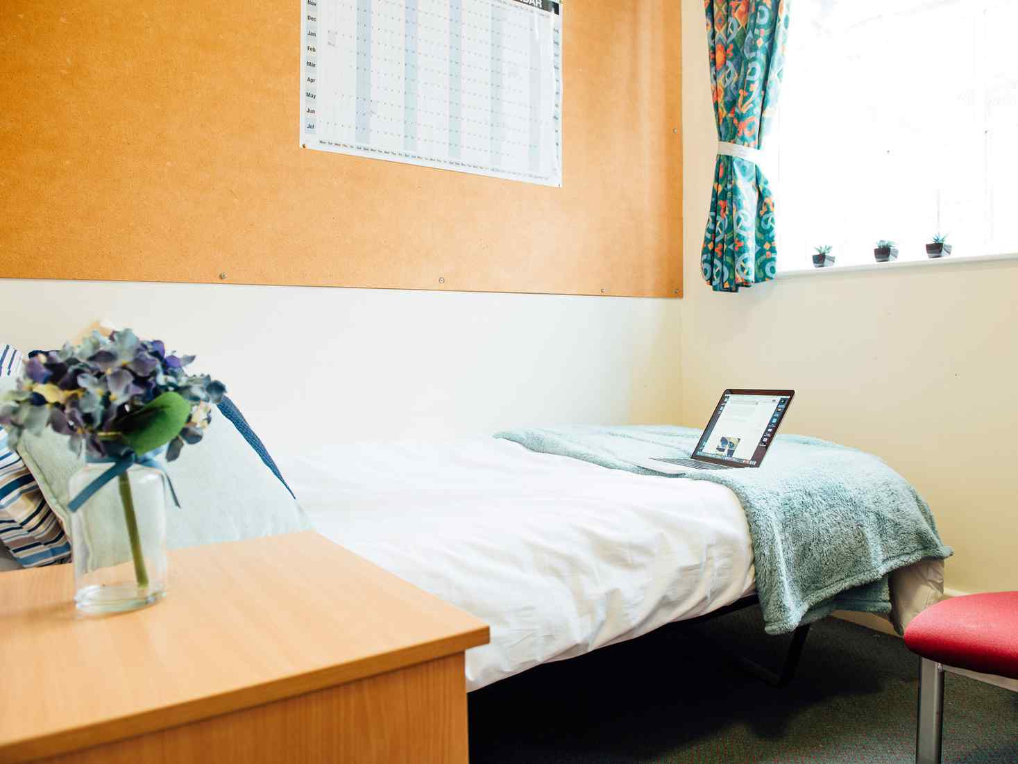 View of a single bed and bedside table in a standard ensuite room at City Residence. A large noticeboard is on the wall above the bed. 