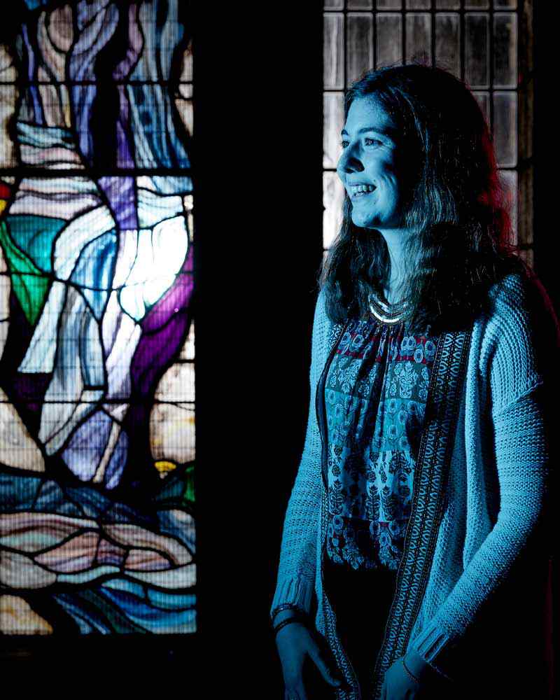 A smiling student stands in front of a stained glass window. 