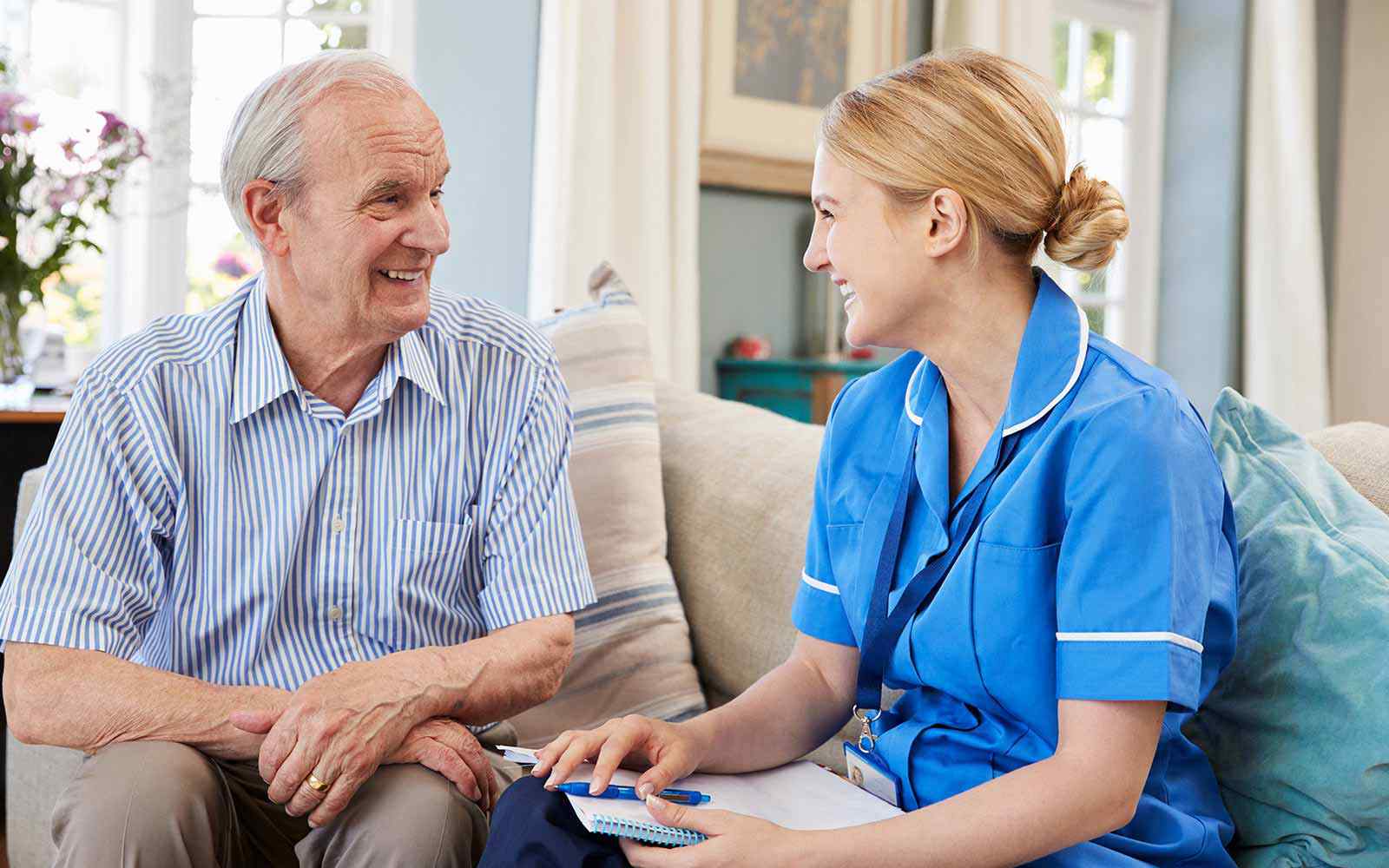 A nurse in a blue uniforms sits and talks with an older patient. 