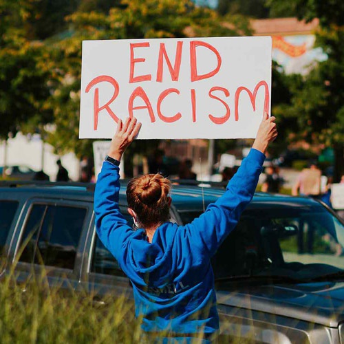 Person in blue jacket outside holding up a sign saying 'End Racism'. 