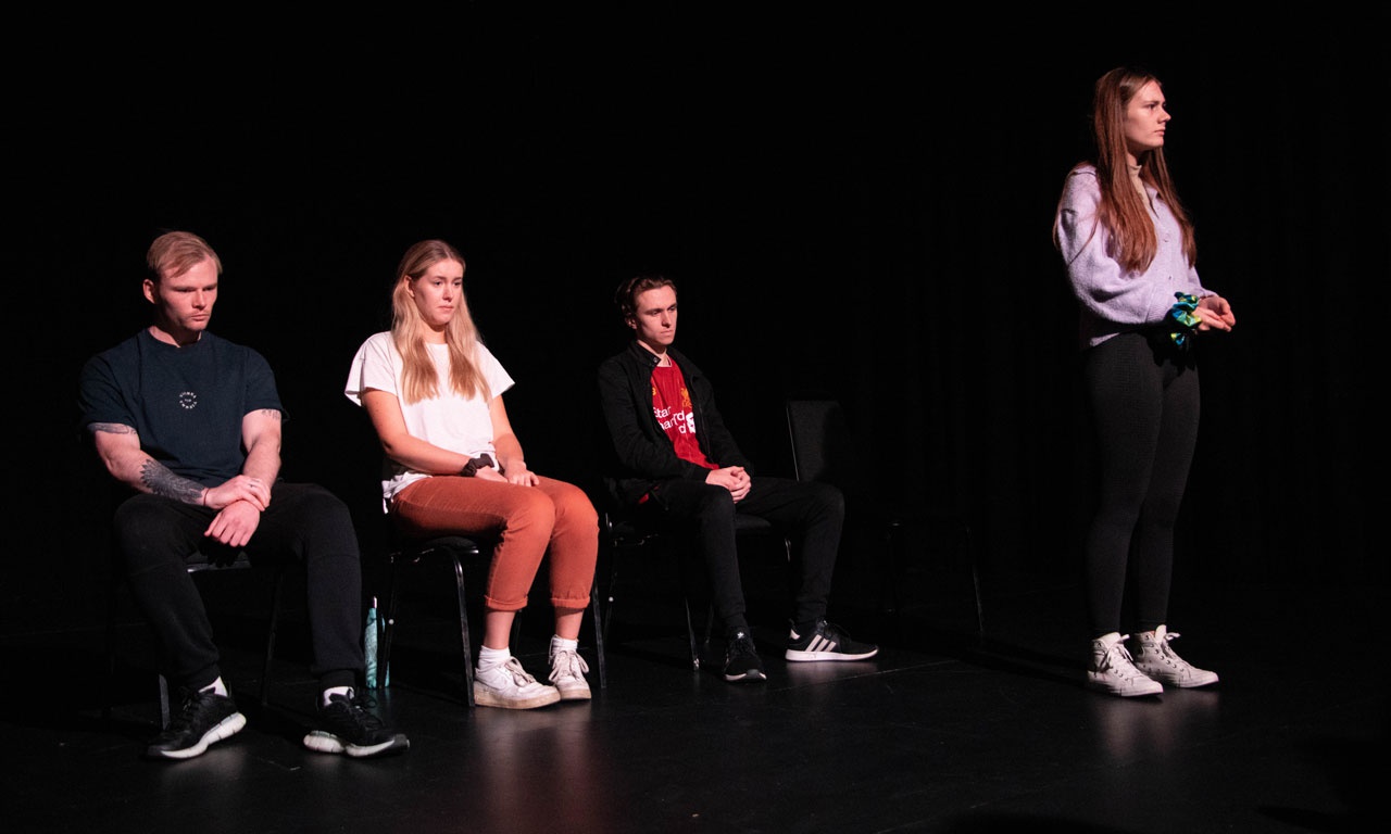 Four actors in Its Not Love play, 3 are sitting on chairs while one stands and addresses the audience. 