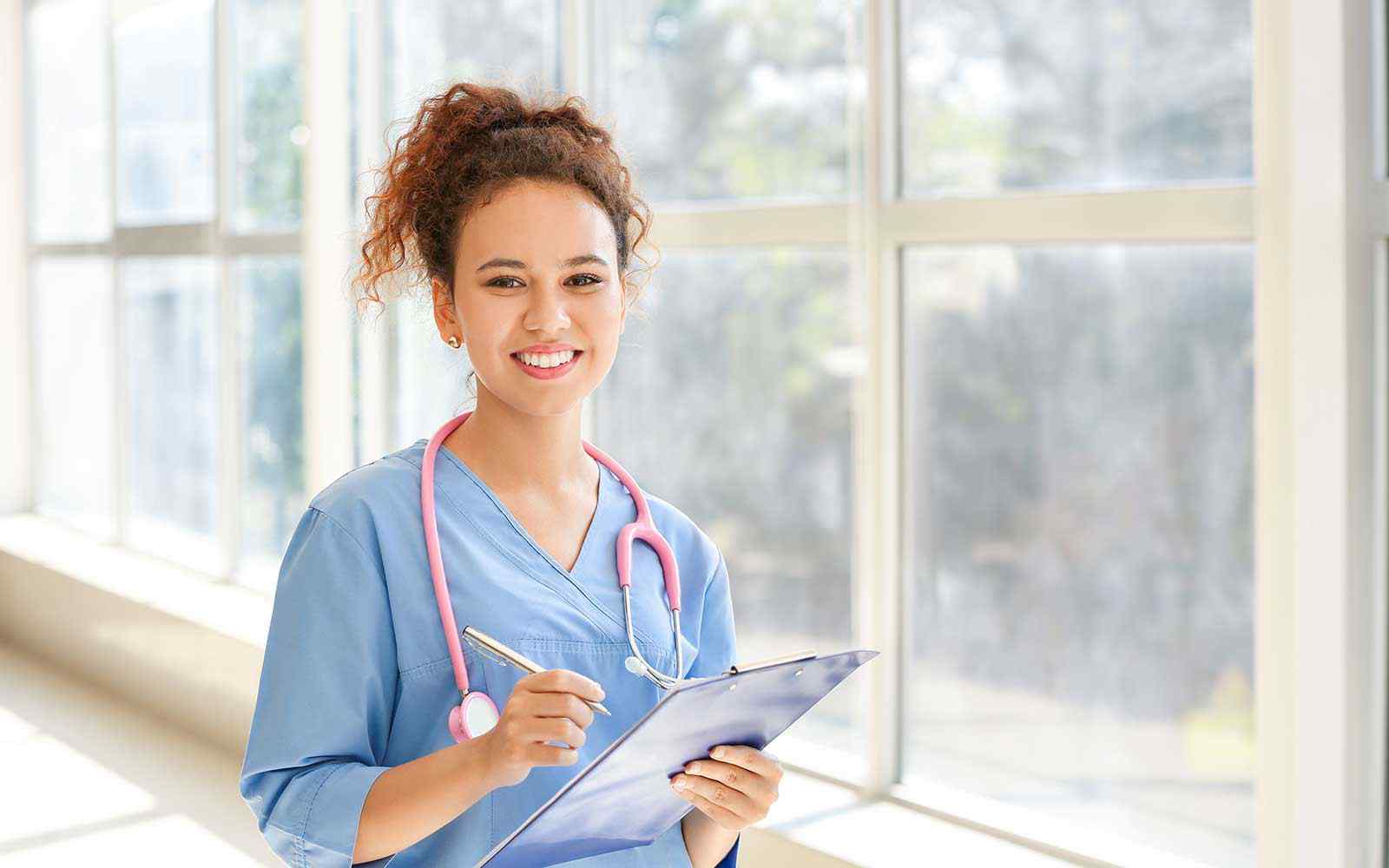 A student in scrubs smiles at the camera. She is holding a clipboard and stethoscope. 