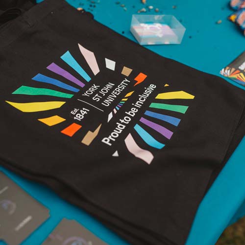 Black tote bag with rainbow sunburst pattern in middle. Underneath the York St John University logo the text reads Proud to be inclusive. 