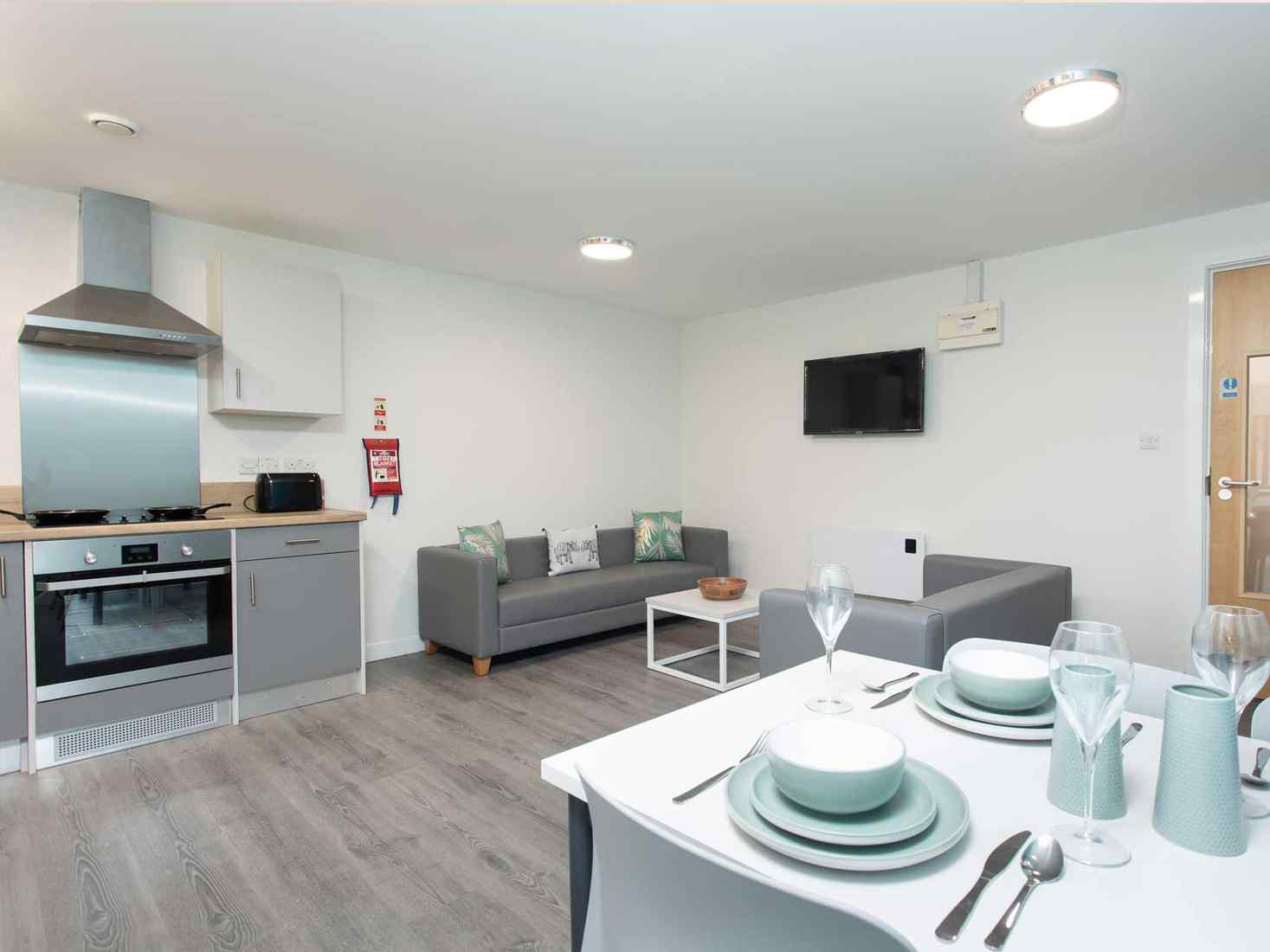 Shared kitchen and living area in Abode accommodation site, with oven, kitchen cupboards, two sofas with TV, and dining table. 