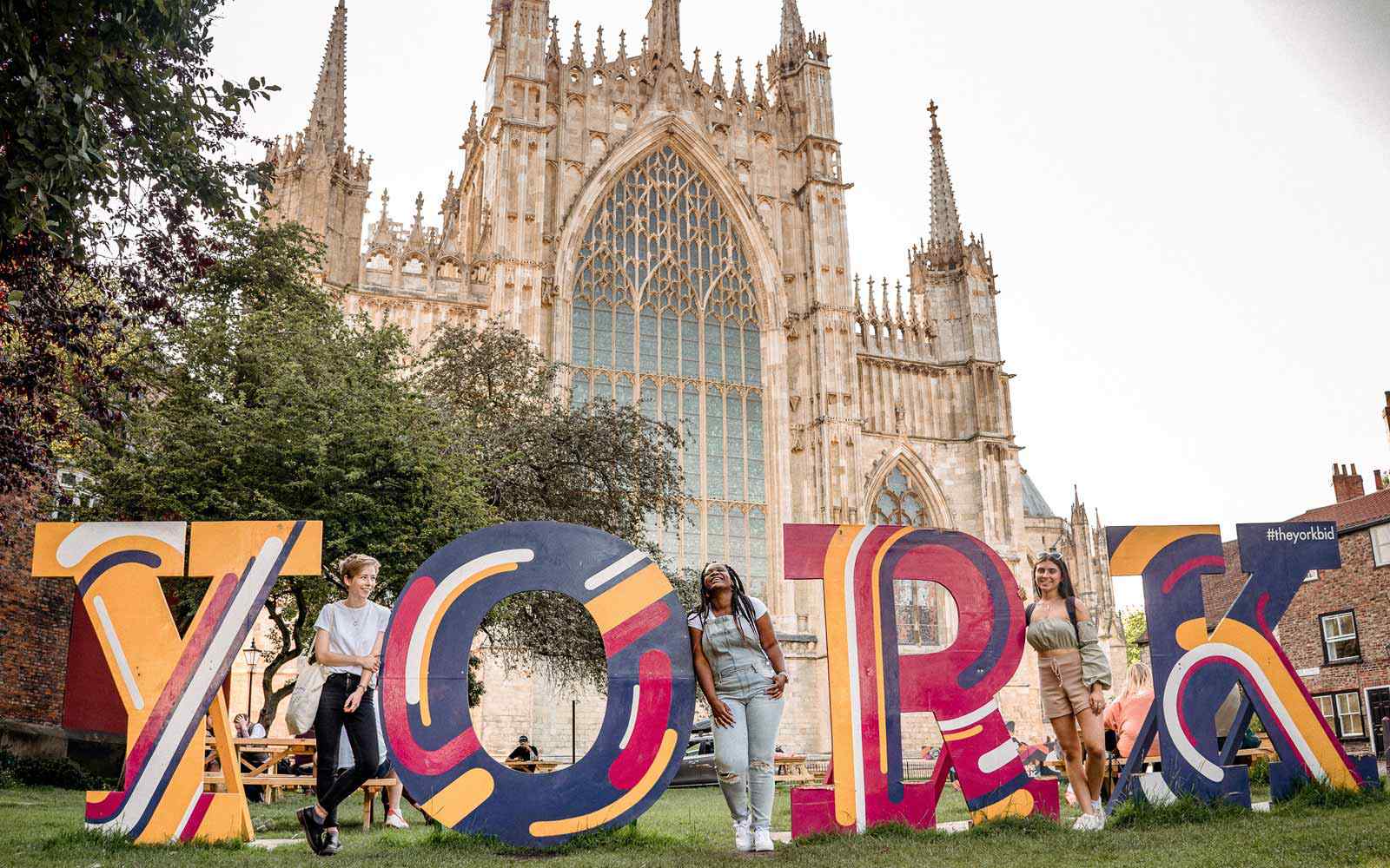 Three students stand in front of York Minster with a giant 'York' sign. 