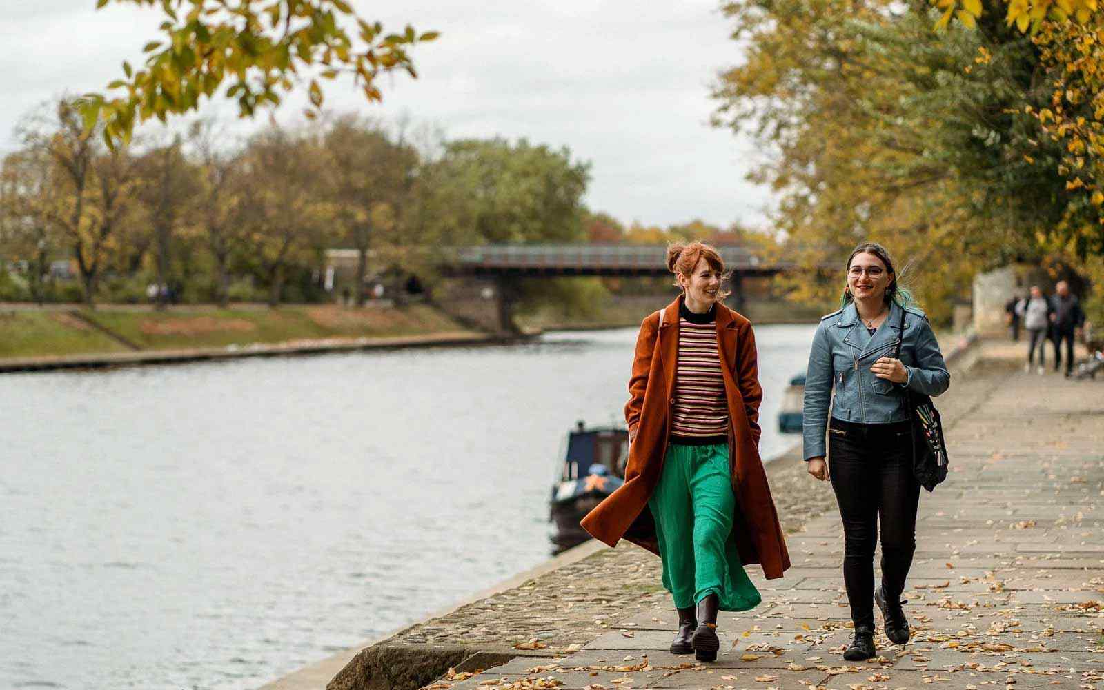 Two students walk together by the river in York, with autumn leaves behind. 